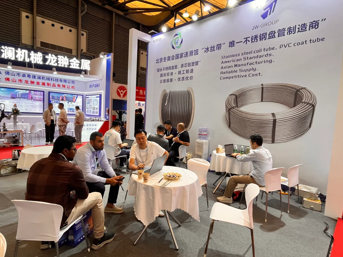 People gathered around discussion tables beside a large stainless steel coil tube display at the JW-GROUP stand